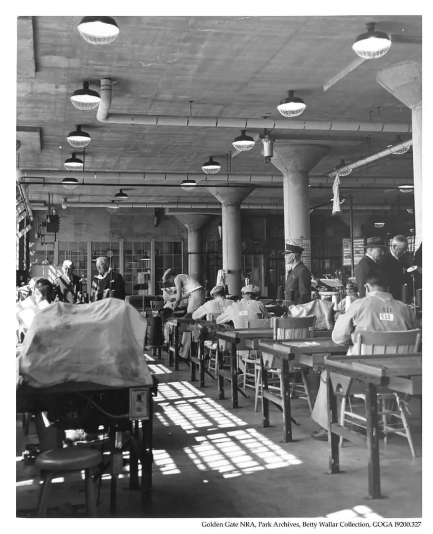Historical black and white photo of Alcatraz prison officials inspecting inmate work inside the prison tailor shop.